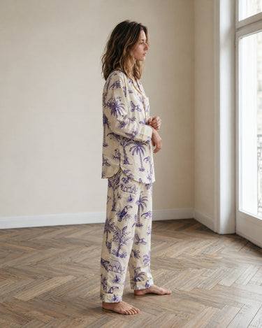 A woman stands in profile wearing a two-piece pajama set with a cream base and purple palm tree print, in a bright room with a wood herringbone floor and a window.