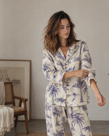 A woman wearing a two-piece pajama set with a blue palm tree and bird pattern on a cream background, standing in a softly lit room.