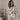 A woman wearing a two-piece pajama set with a blue palm tree and bird pattern on a cream background, standing in a softly lit room.