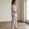 A woman stands in profile wearing a two-piece pajama set with a cream base and purple palm tree print, in a bright room with a wood herringbone floor and a window.