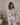 A woman wearing a two-piece pajama set with a blue palm tree and bird pattern on a cream background, standing in a softly lit room.
