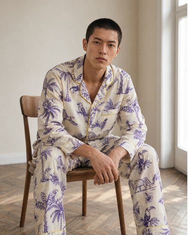 A man sits on a wooden chair wearing a cream-colored pajama set with a purple tropical print of palm trees, birds, and a tiger, in a brightly lit room.