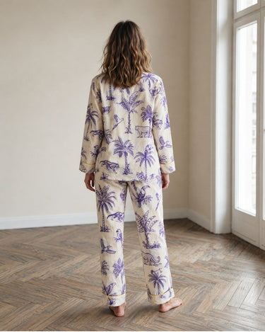 A woman stands with her back to the camera wearing a matching long-sleeve pajama set with a purple palm tree and tropical animal print on a cream-colored fabric, set in a bright room with wooden floors.
